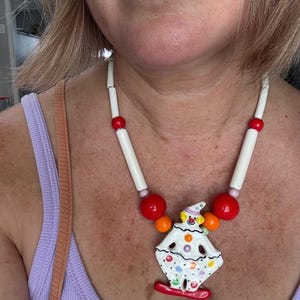May include: A necklace featuring a central ceramic clown pendant with a white body, colorful polka dots, and a red base. The pendant is suspended from a strand of white cylindrical beads, large red spherical beads, and smaller orange beads.