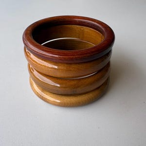May include: Stack of four wooden bangles in varying shades of brown. The top bangle is a deep reddish-brown, while the others transition to lighter, golden hues. The bangles have a smooth, polished finish.