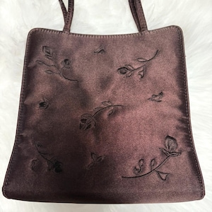 May include: A brown satin tote bag with embroidered floral designs. The bag features a rectangular shape with two thin handles. The embroidered details include roses and leaves, adding a touch of elegance to the accessory.