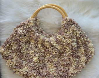 May include: A handmade, textured handbag with a unique design. The bag features a soft, fluffy exterior in shades of beige and mauve, complemented by two wooden handles. The bag's shape is a crescent, perfect for carrying essentials.