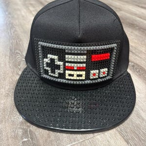 May include: A black baseball cap featuring a flat brim with a textured stud pattern. The front panel displays a rectangular design constructed from small building blocks, mimicking a retro video game controller with a directional pad and buttons.