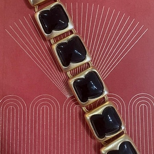 May include: A link bracelet featuring six square-shaped gold-tone metal settings, each containing a smooth, glossy black stone. The links are connected by small, flexible gold-tone metal bars, creating a continuous, articulated chain design.
