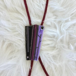 May include: A bolo tie with a rectangular pendant featuring black, purple, and lavender sections. The black section has a zigzag design. The pendant is suspended from a burgundy cord, creating a unique accessory.