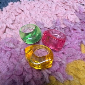 May include: Three chunky, translucent rings in vibrant colors: green, pink, and yellow. The rings are made of a glossy material and sit on a textured, multi-colored surface. The rings have a bold, modern design.