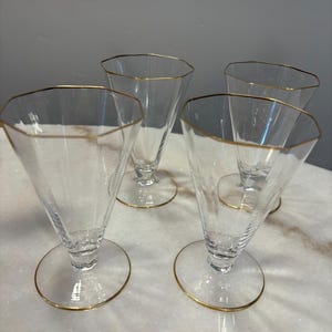 May include: Four clear, vintage champagne glasses with a gold rim and base. The glasses have a unique, faceted design and a conical shape. The glasses are arranged on a white and gray marble surface.