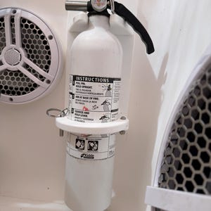 May include: A white fire extinguisher mounted on a white bracket. The extinguisher has a black handle and a pressure gauge. Instructions are printed on the extinguisher. The extinguisher is secured with a metal ring.