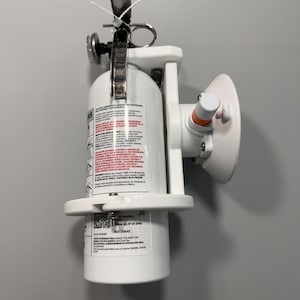 May include: A white fire extinguisher mounted on a white plastic bracket with a silver metal handle and a circular pressure gauge. The cylinder features a label with black text in Spanish, including a serial number 160729646 and safety instructions.