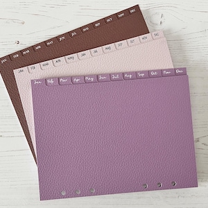 Może przedstawiać: Trzy separatory do planera w odcieniach brązu, różu i fioletu. Każdy separator ma zakładki z miesiącami roku w białym tekście. Separatory mają teksturowany wygląd przypominający skórę i są zaprojektowane z trzema otworami do oprawy.