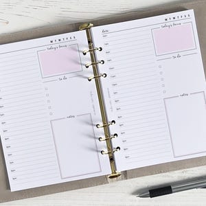 Può includere: Un'agenda giornaliera bianca con un raccoglitore ad anelli dorato. L'agenda ha un programma giornaliero sulla pagina sinistra e una lista di cose da fare e una sezione di note sulla pagina destra. L'agenda ha le parole "today's focus" e "to do" scritte su di essa.
