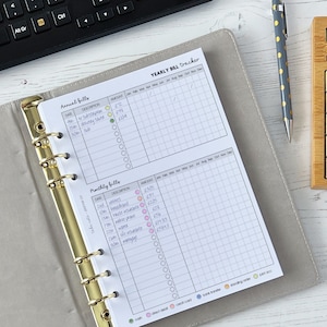 May include: A grey and white planner with a gold binder ring. The planner has a section for "Annual Bills" and "Monthly Bills" with columns for date, description, amount, and check boxes. The planner is on a white wooden surface with a calculator and a pen.