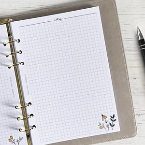 Może przedstawiać: Biały planer z złotym binderem i stroną w kratkę z napisem "notes" u góry. Strona ma mały kwiatowy wzór w prawym dolnym rogu.