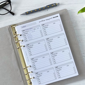 May include: A white purchase repayment tracker with a gold binder ring. The tracker has sections for shop, purchase date, installments, and total. The tracker is on a white wooden surface.