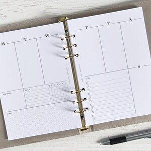 May include: A white planner with a gold binder. The planner has a weekly layout with sections for Monday through Sunday. There are also sections for important tasks, a tracker, and a to-do list.
