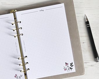 Pode incluir: Um planejador branco com um fichário dourado e páginas pontilhadas. O planejador tem uma seção rotulada como "notas" e um pequeno desenho floral no canto inferior direito.