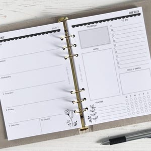 Puede incluir: Agenda abierto con diseño semanal para enero de 2027. Las páginas blancas tienen secciones para prioridades, notas, lista de tareas y un rastreador. Texto negro y elementos decorativos. Un bolígrafo descansa sobre la superficie.