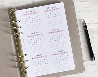 May include: A planner insert with six months of calendars for 2023, in a light pink and white design. The insert is held in a gray binder with gold hardware, and a black pen rests on the side.