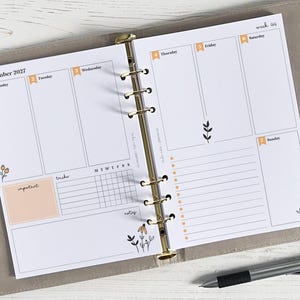 Peut inclure: Un agenda ouvert sur une page de novembre 2027. Les pages blanches sont ornées d'accents orange pour les jours de la semaine et de motifs floraux. Un stylo est posé sur la surface.