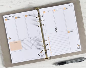 May include: A weekly planner open at a page for November 2027. The white pages have orange accents for the days of the week and floral decorations. A pen lies on the surface.