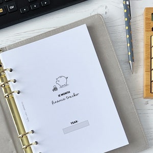 Può includere: Un'agenda bianca con un raccoglitore dorato e un'illustrazione in bianco e nero di un salvadanaio. L'agenda ha il testo "12 MONTH finance tracker" e "YEAR:" sulla copertina.