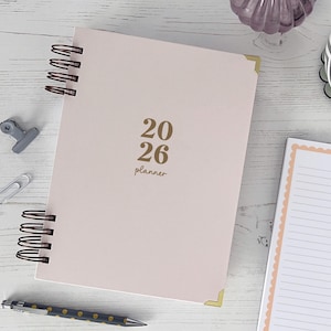 May include: A pale pink 2026 planner with gold lettering and corner protectors. The planner is spiral-bound with black coils. A pen, paper clips, and a binder clip are scattered around the planner on a white wooden surface. A cup of coffee and a decorative glass are also visible.