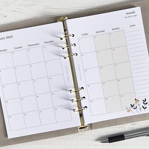 Op de afbeelding: Een plannerinzet voor januari 2027, met een maandelijkse kalenderindeling en een bloemenontwerp in de hoek. De kalender is open in een map met gouden ringen en een pen ligt op het oppervlak.