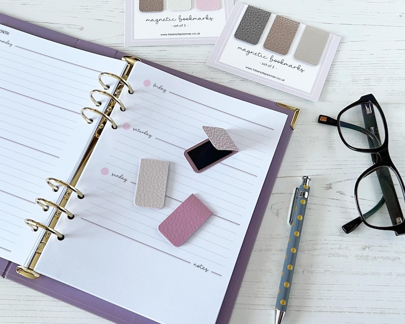 May include: A planner open to a weekly spread with magnetic bookmarks in shades of grey, pink, and beige. A pen and black-framed glasses are also on the white surface. The bookmarks are labelled "magnetic bookmarks - set of 3".