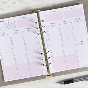 Op de afbeelding: Een open planner met een weekindeling voor november 2027. De pagina's zijn wit met roze accenten en een gouden ringband. De indeling omvat dagelijkse schema's, een wekelijks doelen gedeelte en een "niet vergeten" gedeelte. Een pen ligt op het oppervlak.