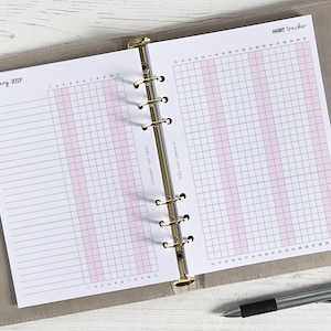 May include: Open planner with habit tracker pages. The left page is labeled "January 2023" and has a grid for tracking habits. The right page is labeled "Habit Tracker" and has a larger grid. A silver pen rests on the bottom right corner.