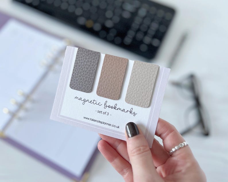 May include: Three magnetic bookmarks in grey, beige and tan, with a textured finish. The bookmarks are displayed on a white card with the text "magnetic bookmarks - set of 3". A planner and keyboard are visible in the background.