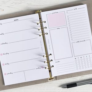 May include: A white planner with a gold binder. The planner has a monthly calendar, a weekly schedule, and a to-do list. It also has a section for notes, priorities, and calls and emails. The planner has a tracker with a grid of circles for each day of the week.