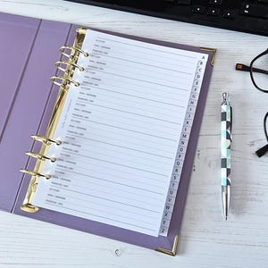 May include: A lavender binder with gold accents, open to a lined page with labeled tabs. A patterned pen and black-framed glasses rest beside it. A keyboard is visible in the background. The binder is designed for organizing information.