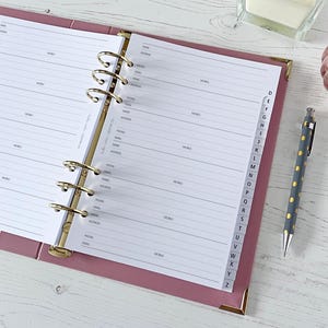 Puede incluir: Carpeta rosa abierta con anillas doradas y páginas de direcciones rayadas. Un bolígrafo gris con lunares dorados descansa al lado. También se ven una bola decorativa de cristal rosa y un pequeño recipiente de cristal.