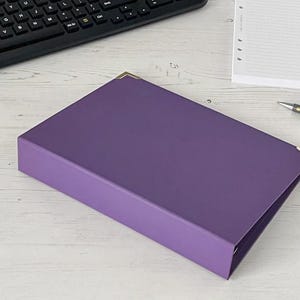 Puede incluir: Un archivador morado con esquinas doradas. El archivador está cerrado y colocado plano sobre una superficie de madera blanca.