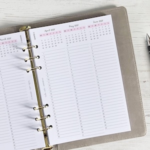 Può includere: Un'agenda con pagine di calendario per marzo, aprile, maggio e giugno 2027, aperta per visualizzare i mesi. L'agenda è in un raccoglitore grigio chiaro con anelli dorati. Una penna nera è appoggiata sul lato destro dell'agenda.