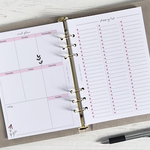 Peut inclure: Un planificateur blanc avec un classeur doré. La page de gauche comporte une section de planification des repas avec les jours de la semaine. La page de droite comporte une section de liste de courses avec une colonne pour chaque jour de la semaine. Le planificateur a un motif floral rose.