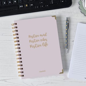 May include: A light pink spiral-bound notebook with gold accents and the words "Positive mind, Positive vibes, Positive life" in gold script. A pen with gold polka dots and a notepad are also visible.