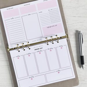 May include: A planner with pink and white pages, open to a January 2027 weekly spread. The planner includes sections for goals, notes, and a tracker. A silver pen rests on the right side of the planner.