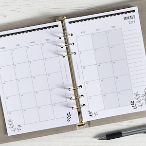 Peut inclure: Agenda ouvert présentant une page de calendrier de janvier 2027. Le calendrier est imprimé sur papier blanc avec une bordure festonnée noire et des motifs floraux. L'agenda est ouvert sur une double page, maintenue par des anneaux dorés, et repose sur une surface gris clair.