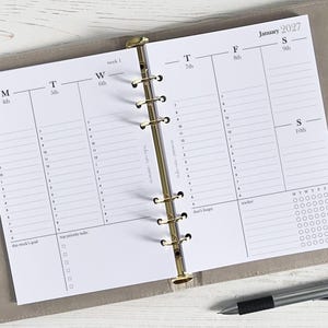 May include: A weekly planner for January 2027, open to a spread with daily schedules, top priority tasks, and a tracker. The pages are white with black text and lines. The planner is held in a light grey binder with a gold-coloured ring binder.