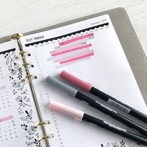 May include: A planner open to a sleep tracker page with pink and gray markers. The sleep tracker is marked with pink highlighting. The planner also has a calendar with floral designs and the text "January 2023".