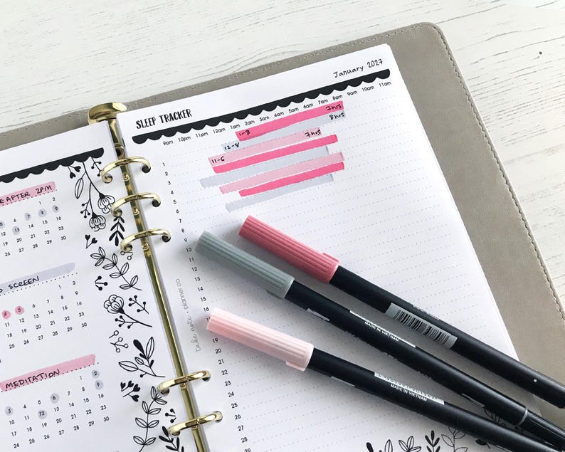 May include: A planner open to a sleep tracker page with pink and gray markers. The sleep tracker is marked with pink highlighting. The planner also has a calendar with floral designs and the text "January 2023".