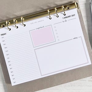 Può includere: Un inserto per planner con un raccoglitore dorato, con un programma giornaliero, una lista di cose da fare e una sezione note. L'inserto include un riquadro rosa per le note importanti e la data "1 gennaio 2027". Una penna d'argento con impugnatura nera è appoggiata accanto al planner.
