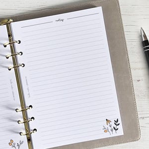 Op de afbeelding: Een witte planner met een gouden binder en gelinieerde pagina's. De planner heeft een sectie met de naam "notes" en een klein bloemenontwerp in de rechterbenedenhoek.
