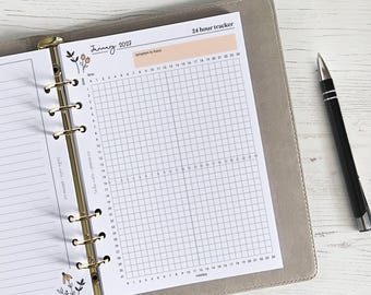 May include: A planner insert with a 24-hour tracker grid, dated January 2023, with space for symptom tracking. The insert is in a gray binder with gold rings. A black pen rests on the right side of the image.