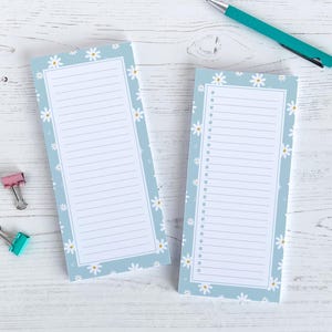 May include: Two rectangular notepads with a light blue border and daisy flower pattern. The notepads have lined pages for lists and a check box column. A teal pen and two binder clips are also visible.