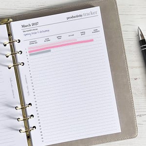 May include: A productivity tracker planner page for March 2027, with a focus on spring cleaning and decluttering. The page is open in a gray binder with gold rings, and a black pen rests on the side. The tracker has pink and gray progress bars.
