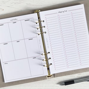 Peut inclure: Un planificateur blanc avec un classeur doré. Le côté gauche comporte une section de plan de repas avec les jours de la semaine étiquetés. Le côté droit comporte une section de liste de courses avec des lignes pour écrire.