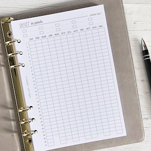 May include: A planner page for the year 2027, featuring a grid layout for tracking. The page is inside a gray binder with gold accents. A black pen rests on the side. The text "2027 in pixels" is visible at the top.