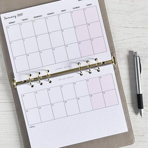 Peut inclure: Un planificateur ouvert sur une page de calendrier de janvier 2017. Le calendrier est dans un classeur gris avec des anneaux dorés. Un stylo argenté avec une prise en main noire repose sur le côté droit du planificateur. Le calendrier a des surlignages roses.