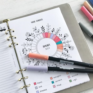 May include: A planner open to a "Mood Tracker" page for January 2024, with a floral design and colour-coded sections. Several coloured markers are placed around the planner. The planner is open to a lined page and a mood tracker page.